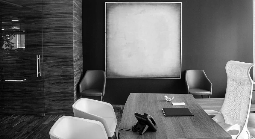 Modern office conference room with a wooden table, white and dark chairs, a telephone, and a large blank square painting on the wall.