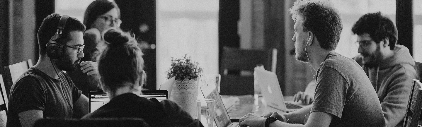 Group of five people working together at a table with laptops and headphones in a collaborative workspace.