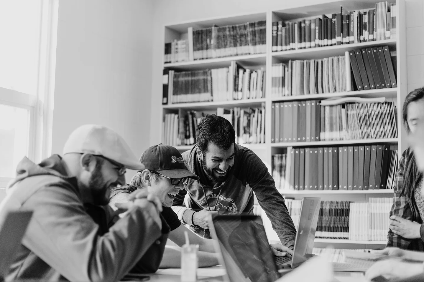 Group of young adults laughing together around a laptop in a library setting.