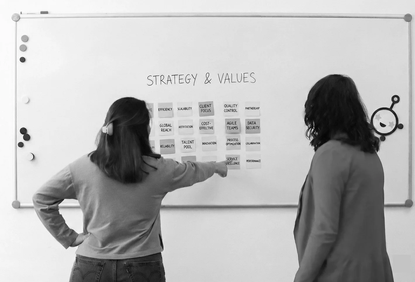 Two women discuss sticky notes on a whiteboard titled 'Strategy & Values' with words like efficiency, client focus, and data security.