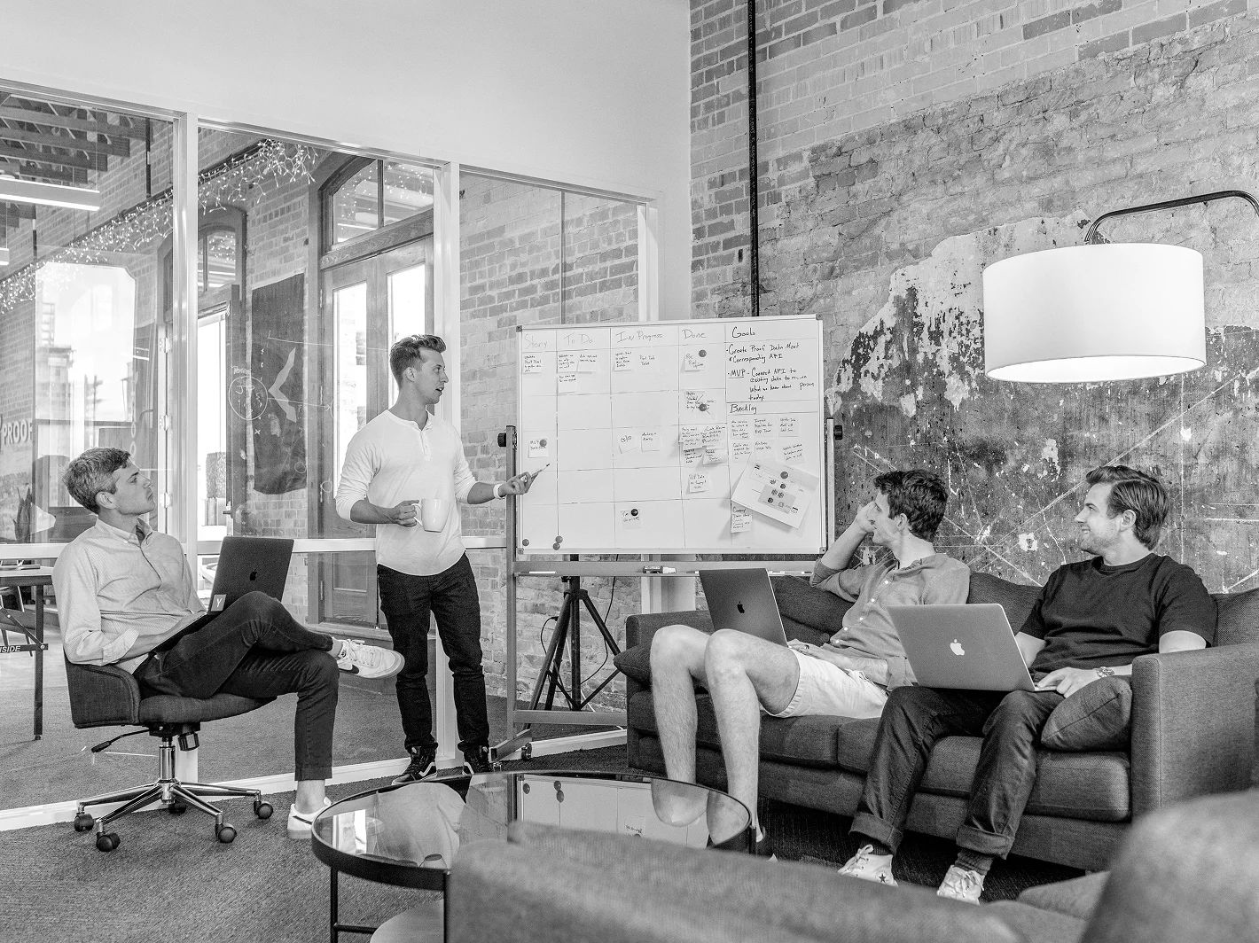 Four men in a modern office having a meeting with one standing and pointing at a whiteboard while others sit with laptops.
